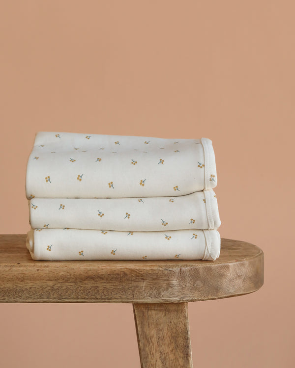 Three neatly folded Organic Baby Blanket – Gold Sprig, featuring a cream color with a delicate floral print, are stacked on a wooden bench against a plain peach background.