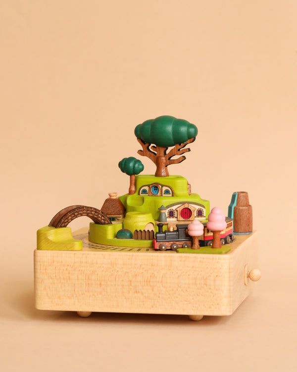 A whimsical miniature Wooden Train Music Box - Spring depicting a vibrant scene with tiny, detailed trees, a bridge, and buildings against a plain beige background. This hand-cranked music box is crafted from sustainably