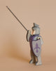 A charming Ostheimer Blue Knight - Riding, meticulously handcrafted from wood, poses with its right arm raised, clutching a long spear. The knight wears a shimmering silver suit of armor and carries a shield adorned with a purple fleur-de-lis emblem. Against a simple beige backdrop, it creates an ideal setting for imaginative play.