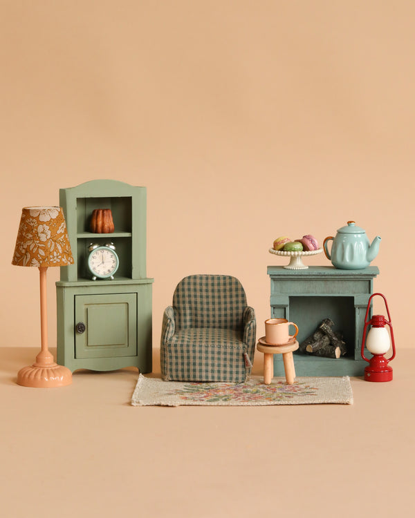 The Maileg Living Room Set features a miniature scene with a lamp, cabinet and clock, armchair, side table with Miniature Tea Set and pastries, pink mug, rug, fireplace, and red lantern—all arranged against a beige background.