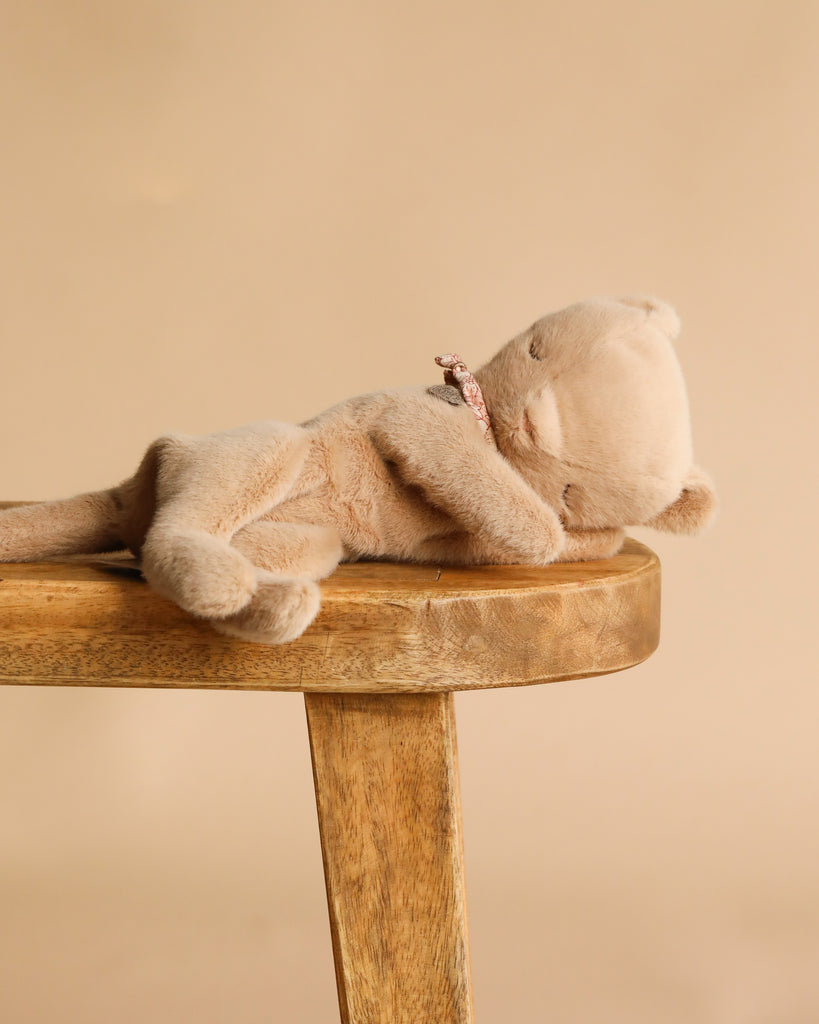 The Maileg Sleeping Kitten Plush, Small - Cream Peach, with a pink collar, lies on its side atop a wooden stool against a plain beige background.