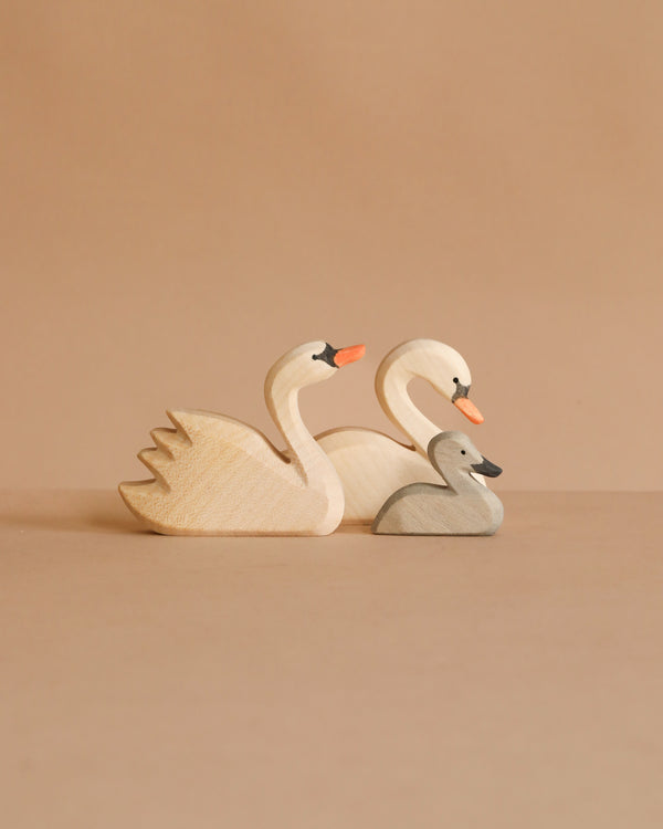 The Ostheimer Handmade Wooden Swan Family Set features two large swans with orange beaks and a small gray cygnet, all handcrafted from wood and arranged on a beige background.