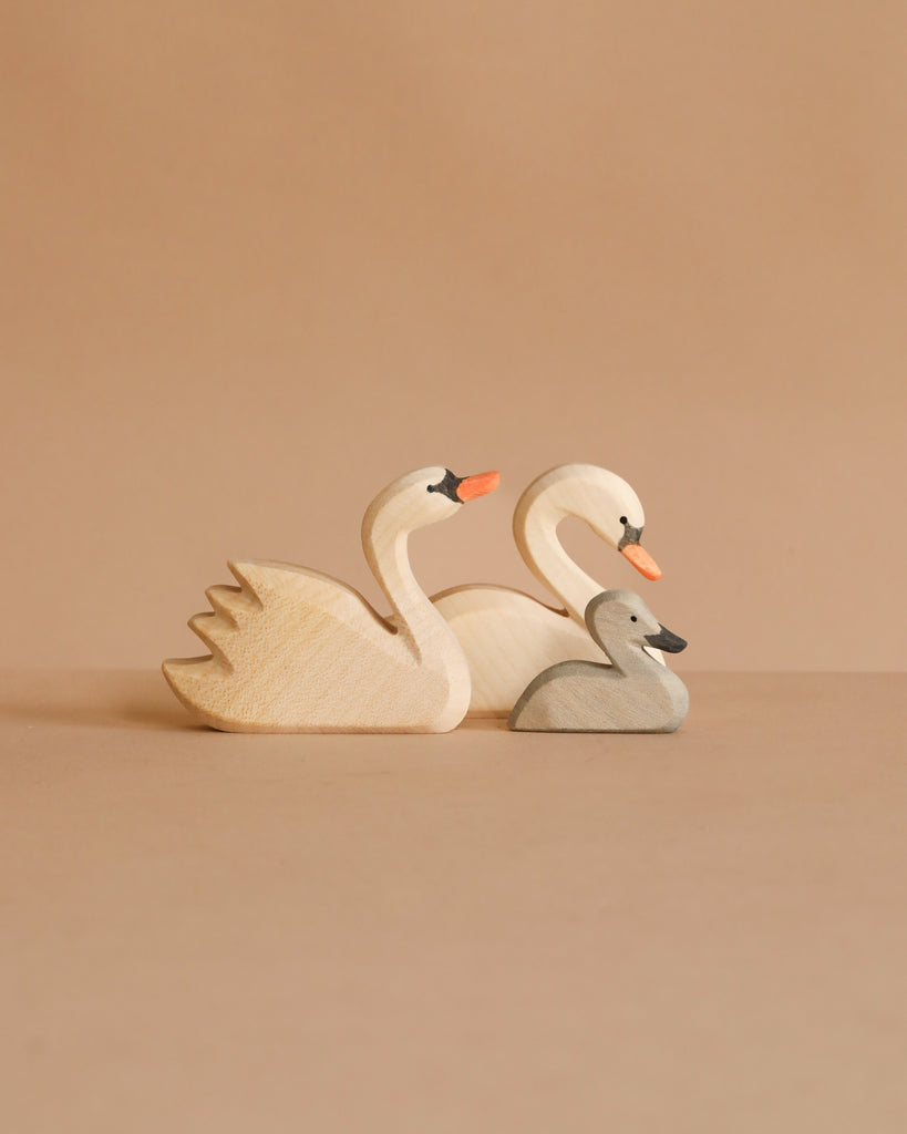 The Ostheimer Handmade Wooden Swan Family Set features two large swans with orange beaks and a small gray cygnet, all handcrafted from wood and arranged on a beige background.