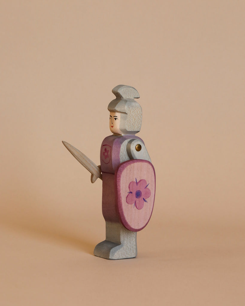 An Ostheimer Red Knight - Standing, a small wooden toy figurine, stands against a plain beige background. The knight, handcrafted from sustainably sourced materials, is adorned with a silver helmet and a lavender chest plate while wielding a sword. Holding a pink shield that features a flower emblem, the knight is viewed in side profile looking left.