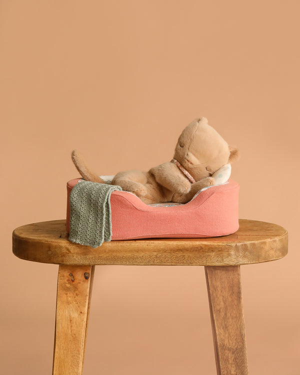 The Maileg Small Sleeping Kitten in Cozy Bed lies on its back in a pink pet bed with a green knit blanket, set on a wooden stool against a beige background.