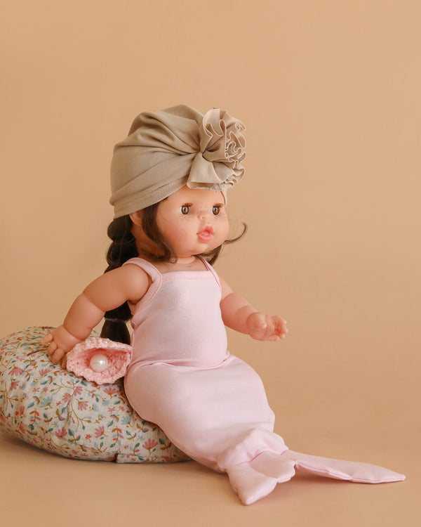 The Minikane Baby Doll (13") - Valentine wears a light pink mermaid costume and beige turban, seated on a floral cushion with a small pink crochet bag beside it, set against a beige background.