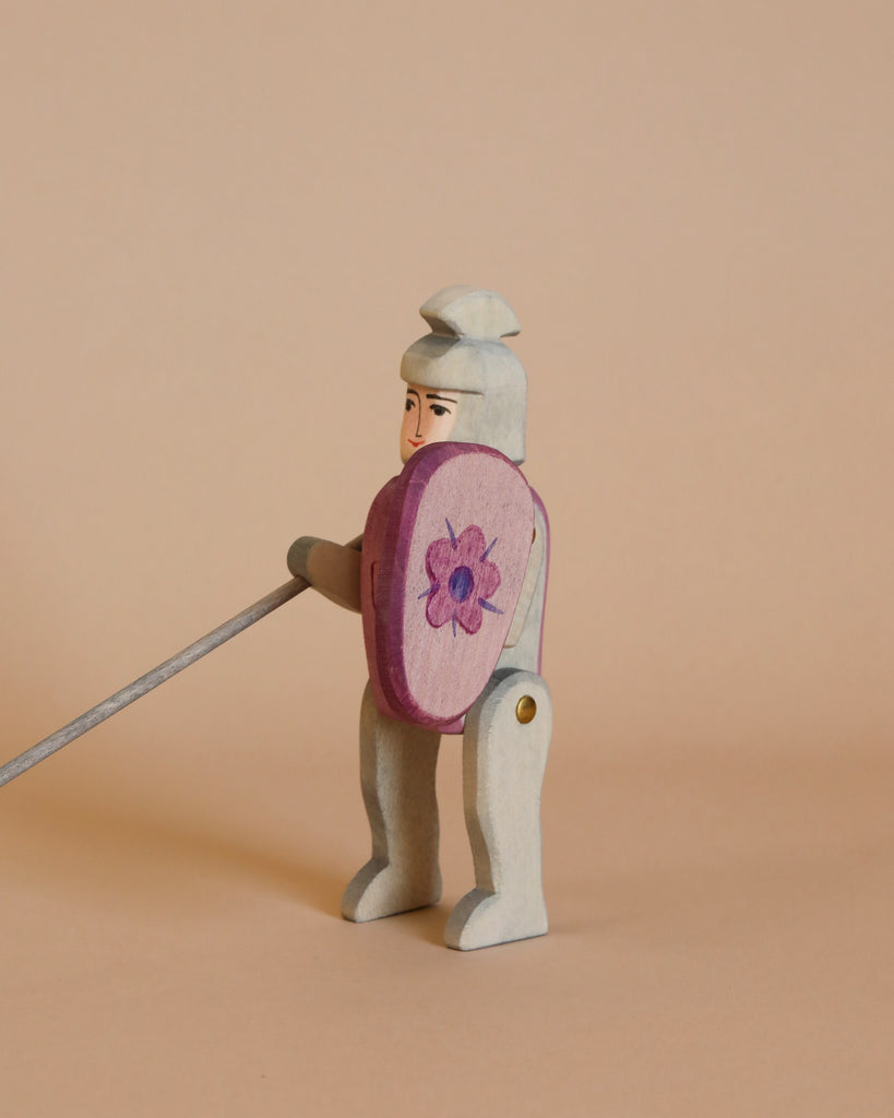 An Ostheimer Red Knight figure stands against a beige background. The handcrafted wooden toy, part of the Ostheimer toys collection, holds a long sword in one hand and a shield painted with a pink flower in the other. Adorned with a simple white helmet and armor, it inspires imaginative play.