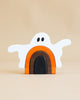 A Handmade Wooden Ghost Stacker depicting a white ghost with outstretched arms and black eyes, standing over an arch made of three stacked shapes in orange, brown, and black. Handmade from poplar hardwood and finished with a non-toxic wood dye, the background is a neutral beige color.