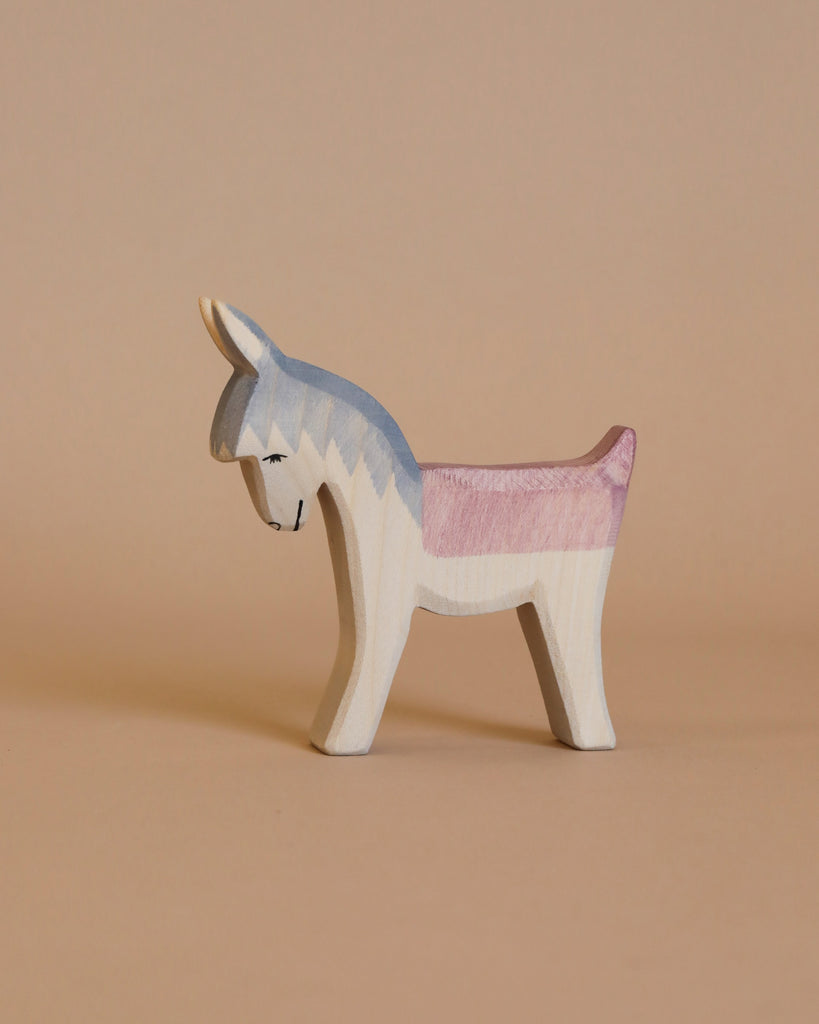 An Ostheimer Mary on Donkey handcrafted wooden toy features a painted color scheme resembling the transgender pride flag, with light blue, pink, and white hues. Perfect for imaginative play, this charming toy is set against a plain, light beige background.