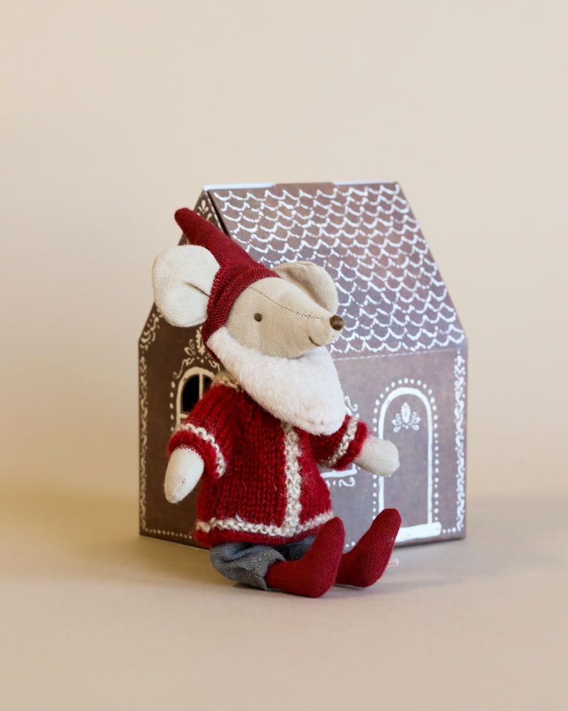 A Maileg Santa Mouse in a red outfit and hat sits in front of the Maileg Gingerbread House Christmas Set box, decorated with white icing-like designs to resemble a festive gingerbread house, all against a beige background.