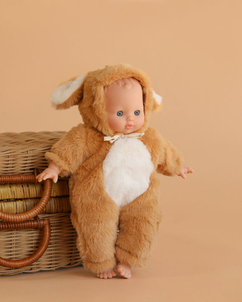 The Minikane Soft Body Doll (11") - Claire with Bambi Costume, featuring a fuzzy brown and white outfit with floppy ears, stands beside a wicker basket on a beige background.