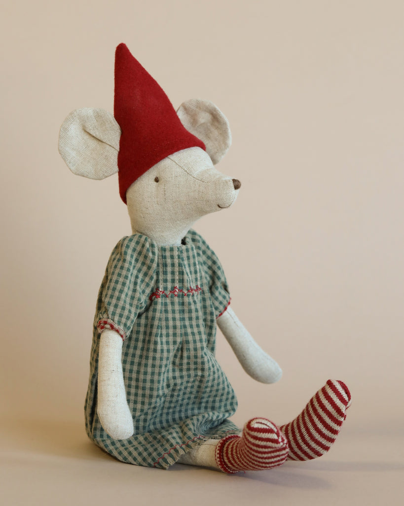 A Maileg Medium Christmas Mouse - Girl dressed in a green plaid jumpsuit and striped socks, wearing a red pointed hat, sitting against a neutral background.