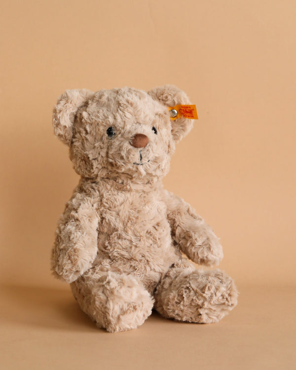 A Steiff, Honey Teddy Bear, 11 Inches with shaggy, light brown fur sits against a plain beige background. The bear has friendly black eyes, a stitched nose, and a small, rectangular "Button in Ear.