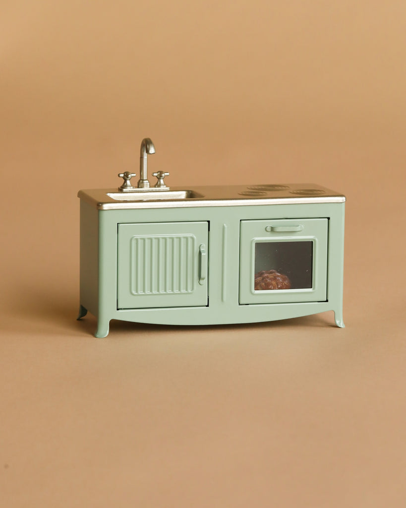 The Maileg Kitchen Set with Chef Mouse, part of the mouse playset collection, is a small mint green kitchen with a sink, cabinet, oven containing a toy item—perfect for miniature food accessories—shown on a beige background.