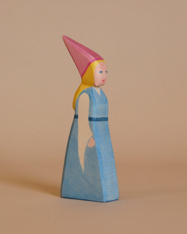 Introducing the Ostheimer Daughter of the Castle: a handcrafted wooden figurine of a woman adorned in a blue dress and a tall pink hat. This charming figure features yellow hair, simplistic facial details, and is accented with a light blue sash around her waist. Made from sustainably sourced materials, she stands elegantly against a plain light brown background.