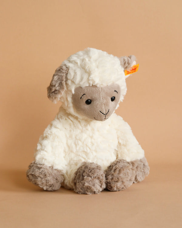 A Steiff, Lita Lamb Plush Animal Toy, 12 Inches sitting against a beige background, featuring fluffy cream-colored wool, with a beige face and limbs, and a small smiling expression.