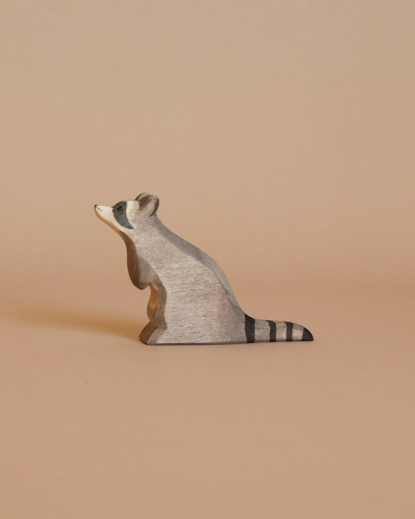A small, handcrafted wooden raccoon with a gray body and black-striped tail from the Ostheimer Handmade Wooden Woodland Animals Set sits on a plain, light brown background, capturing the charm of Ostheimer toys.