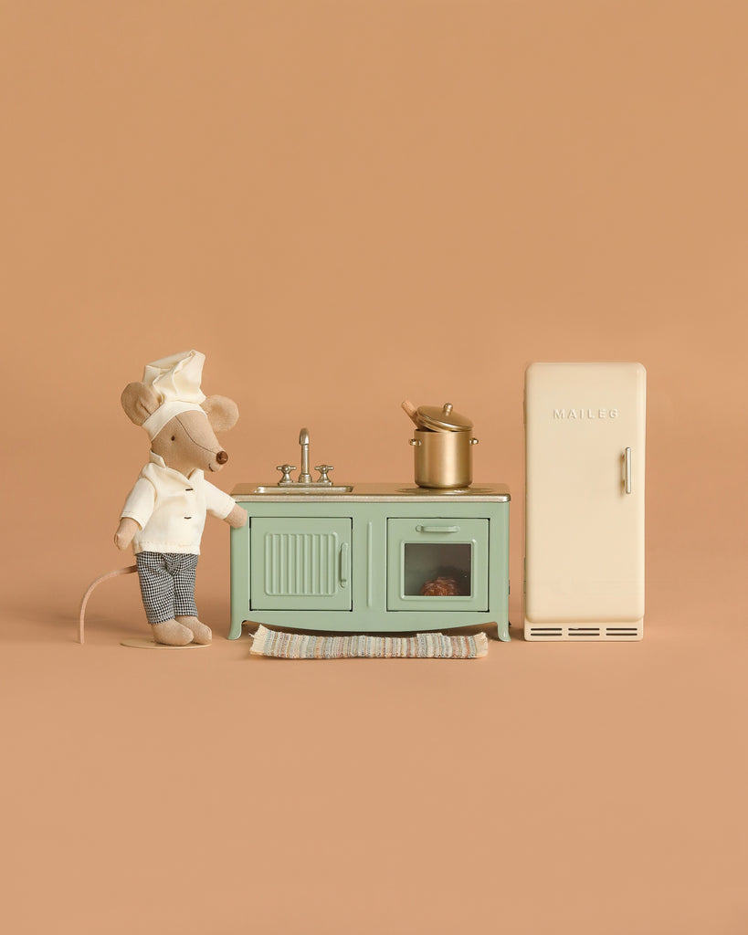 The Maileg Kitchen Set with Chef Mouse features a toy mouse chef beside a mint green stove and sink, with a pot on the stove and a beige refrigerator, all set against a plain beige background.