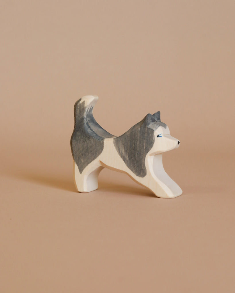 The Ostheimer Sled Dog - Running is a small, wooden figurine with a playful stance, painted in white and gray hues. This handcrafted piece features its front legs extended forward and its tail raised, capturing a stretching or playful pose. The background is a plain, light brown color.