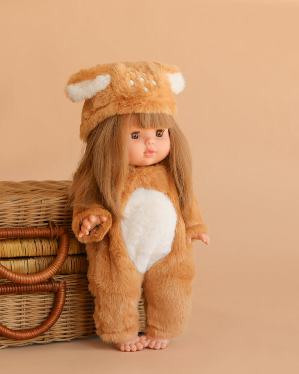 The Minikane Baby Doll (13") - Nina with Bambi Costume, featuring long brown hair and a fuzzy brown outfit with a white belly and matching hat with ears, stands beside woven baskets against a beige background.