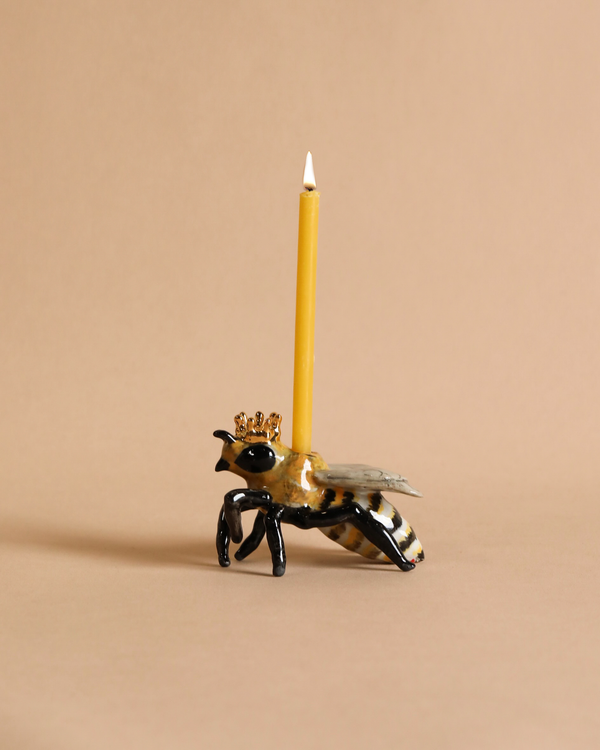 A whimsical candle holder shaped like a Queen Bee Cake Topper, with a lit yellow candle positioned upright on its back, against a soft beige background. This fine porcelain piece highlights delicate artistry and charm.