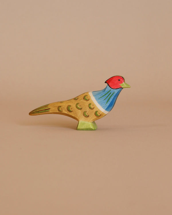 The Ostheimer Pheasant is a handcrafted wooden bird figurine made from sustainably sourced materials, featuring colorful painted details such as a red head, blue neck, and yellow and brown body, displayed against a plain beige background.