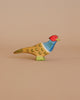 The Ostheimer Pheasant is a handcrafted wooden bird figurine made from sustainably sourced materials, featuring colorful painted details such as a red head, blue neck, and yellow and brown body, displayed against a plain beige background.