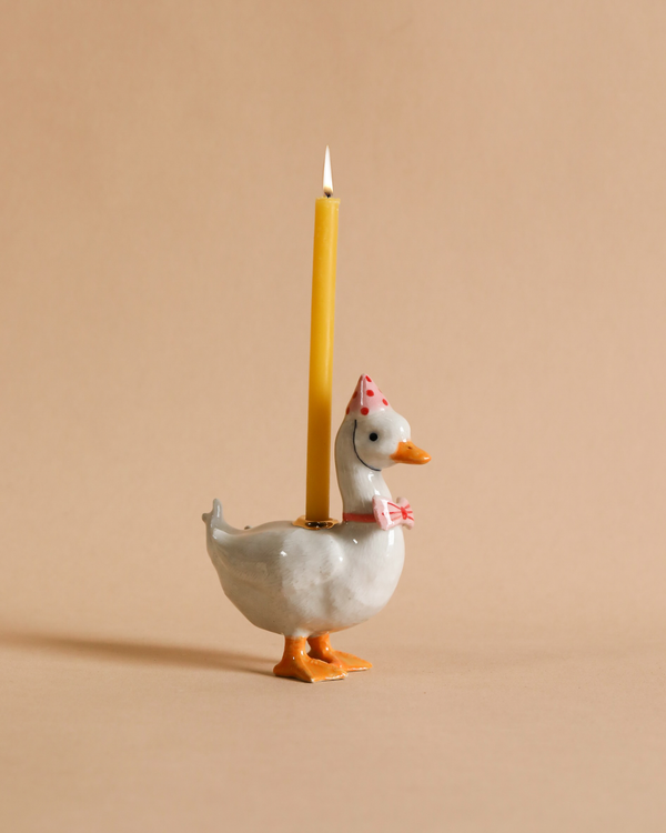 A hand-painted ceramic Pink Goose Cake Topper wearing a red polka dot bonnet and a matching bow tie, standing upright with a yellow candle attached to its back, lit against a plain beige background.