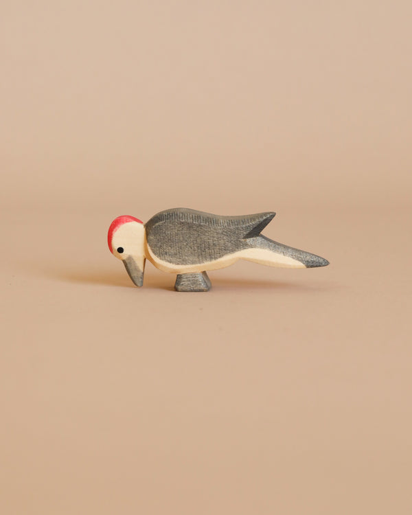 The Ostheimer Woodpecker, a small wooden bird figurine with a red head and gray wings, stands gracefully on a beige surface. Crafted in a minimalistic style with smooth lines and muted colors, this piece exemplifies the exquisite use of sustainably sourced materials often found in handcrafted wooden toys.