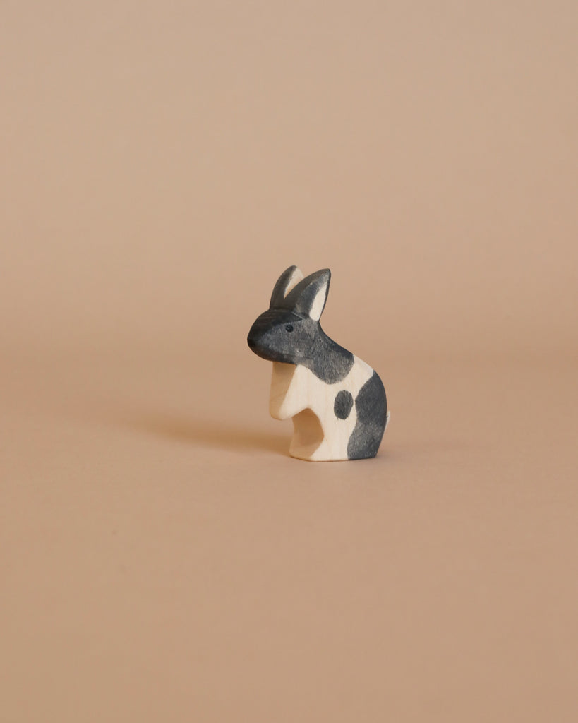 A small handmade wooden rabbit from the Enchanted Woodland Farm Set, featuring black and white patches, sits on a light beige surface with a matching background, evoking the charm of Ostheimer animal figures.