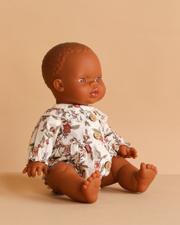The Minikane Doll (13") - Zélie With Clothing, a brown-skinned doll made in Spain, is shown upright in a white floral outfit on a beige surface, facing slightly right against a plain beige background.
