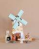 A Penny Windmill set against a beige background, accompanied by small wooden animal figures, a fireplace, and cooking utensils. The scene evokes a rustic, playful ambiance suitable for imaginative play.