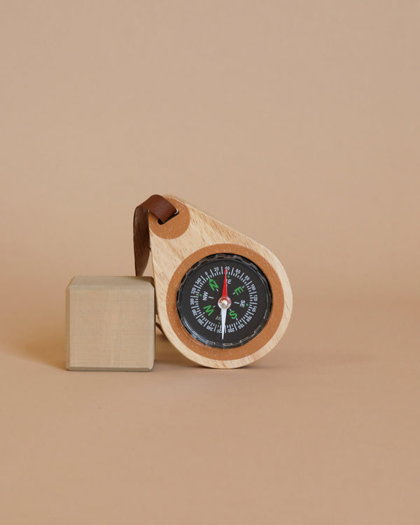 A Wooden Compass, ideal for young adventurers, with a leather strap partially rests against a small wooden cube on a neutral beige background. The compass boasts a black face with cardinal directions and markings, igniting children’s imaginative play.