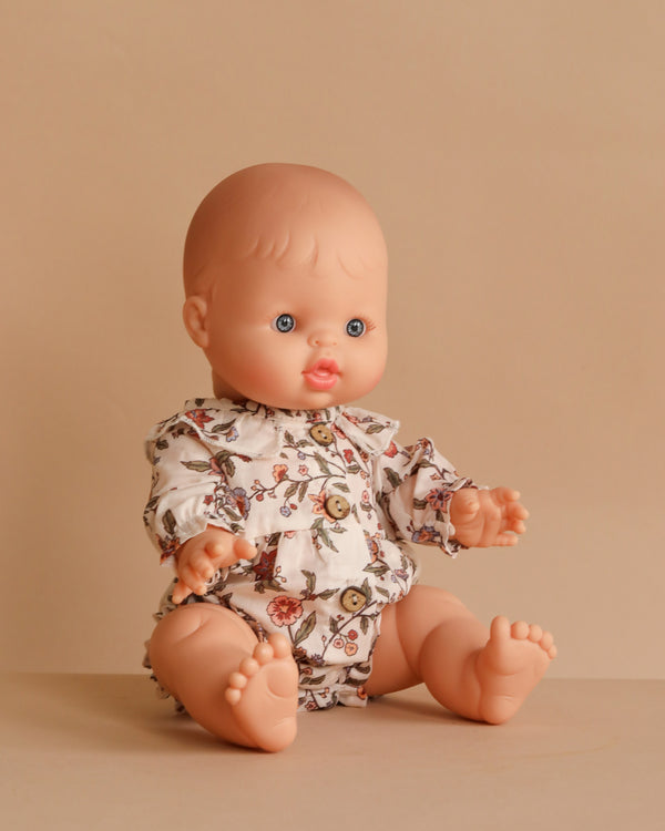 The Minikane Doll (13") - Lily With Clothing, featuring blue eyes and a floral romper, sits upright on a beige surface with a matching beige background.