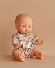The Minikane Doll (13") - Lily With Clothing, featuring blue eyes and a floral romper, sits upright on a beige surface with a matching beige background.