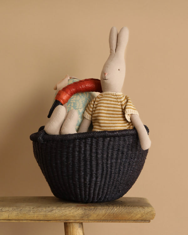 The Easter Basket Set features a stuffed rabbit in a striped shirt with a plush sausage, nestled in a handmade Bolgatanga basket, on a wooden table against a beige backdrop.