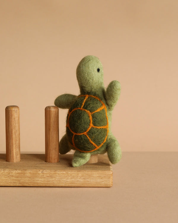 The Turtle Finger Puppet features a green felt body and an orange-outlined shell, standing upright on a wooden base with two dowels. This charming piece is handcrafted by skilled artisans in Nepal.