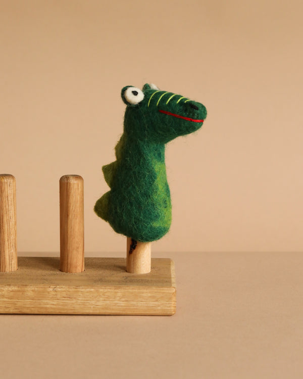 The Felt Gator Finger Puppet, featuring big eyes and a green felt design, is displayed on a wooden stand with three pegs against a beige background—ideal for imaginative play and storytelling.
