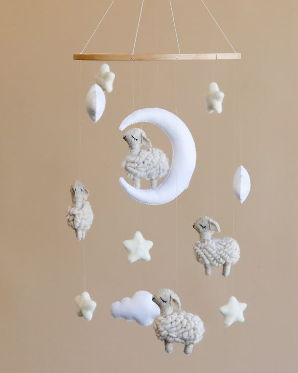 A gentle nursery mobile featuring a crescent moon and several fluffy sheep, along with small white stars, all hanging from a simple wooden frame against a soft beige background.