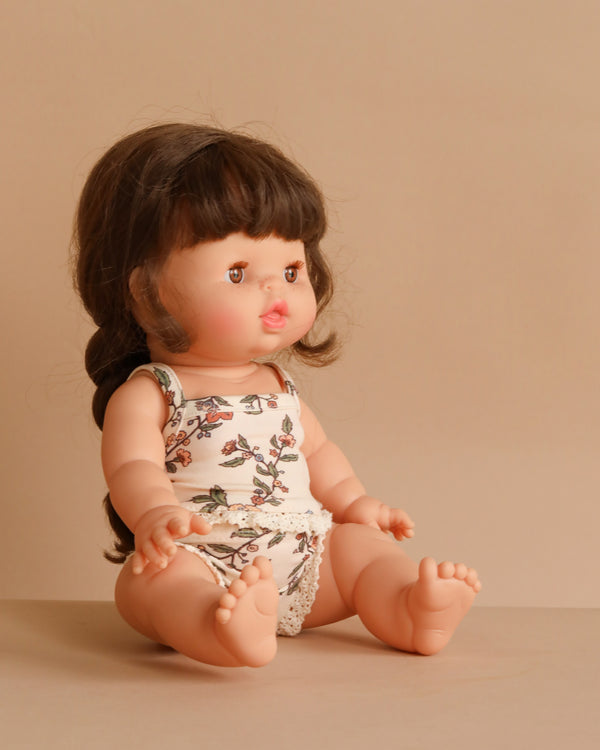 The Minikane Doll (13") - Valentine With Clothing features brown hair in pigtails and wears a floral-patterned white tank top with matching underwear. This anatomically correct doll, made in Spain, sits on a neutral surface with a beige background.