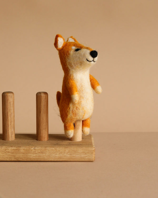 The Shiba-Inu Dog Finger Puppet, a small handmade wool figure in orange and white, stands upright on a wooden pegboard with a beige background.