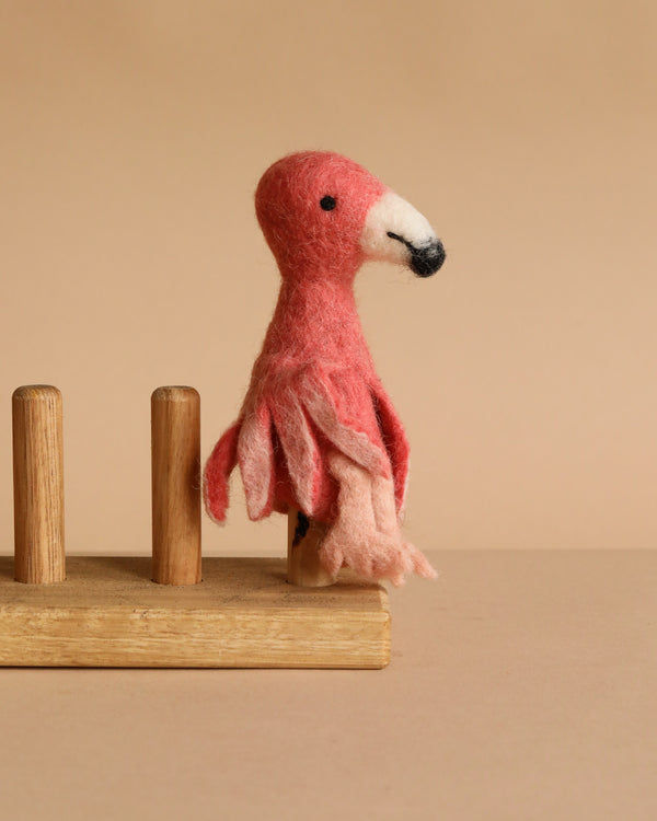 The Flamingo Finger Puppet, handmade with pink and light pink felted details, sits on a wooden stand against a beige background, inspiring imaginative play.