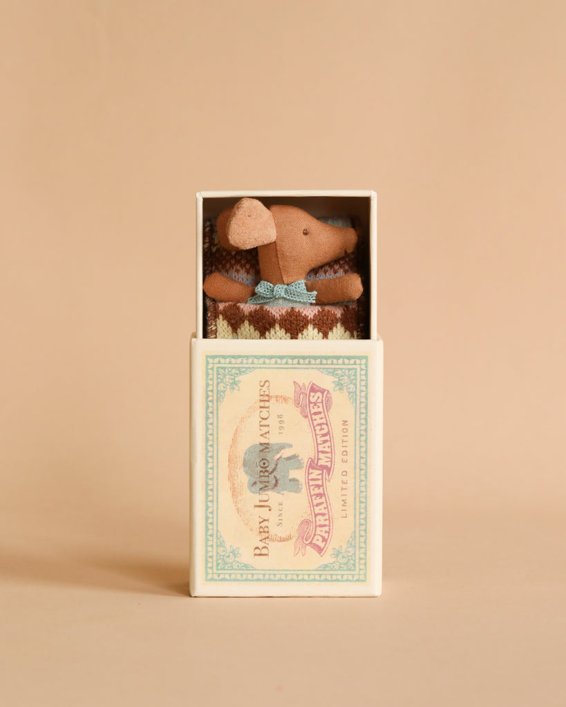 A Maileg Sleepy Wakey Baby Mouse In Matchbox - Blue with long ears is tucked neatly inside a small cream-colored matchbox. The matchbox features a vintage-style label with the text "Bear Twin Matches" and an illustration of an elephant. The background is a warm beige color.