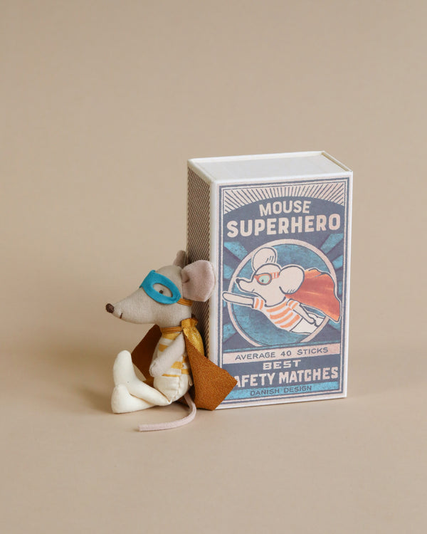 A Maileg | Superhero Mouse, Little Brother figurine, dressed as a superhero, wearing a mask and cape, is leaning against a colorful matchbox labeled "Little Brother Superhero" on a beige background.