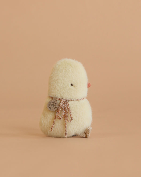 The Maileg Chicken Plush, Mini is a small yellow chick stuffed toy wearing a brown ribbon and round tag, shown sitting away on a peach background.