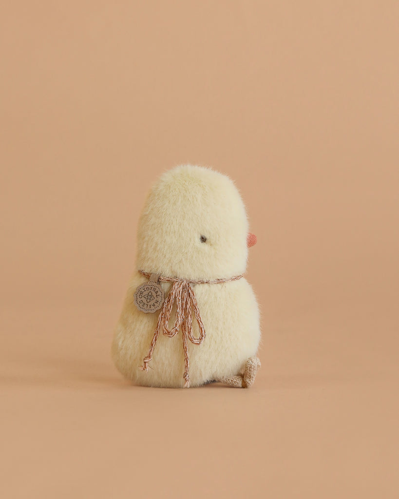 The Maileg Chicken Plush, Mini is a small yellow chick stuffed toy wearing a brown ribbon and round tag, shown sitting away on a peach background.