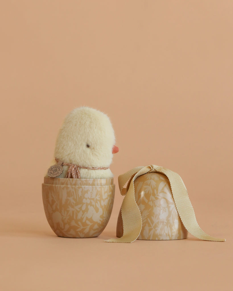 The Maileg Egg With Mini Chick features a soft yellow plush chick inside a decorative beige egg-shaped container with a floral pattern. The lid, adorned with a ribbon, sits beside the set on a matching peach background.