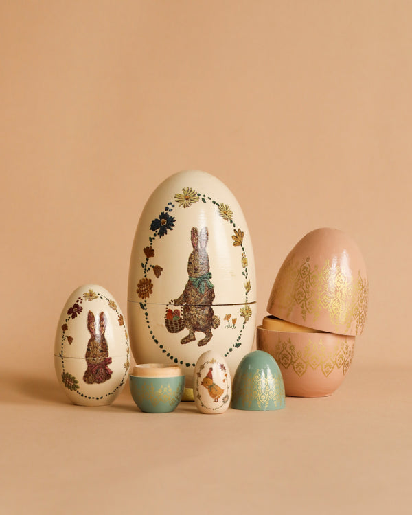 A collection of intricately painted Maileg Easter Babushka Eggs, including a large Easter babushka egg with a floral rabbit design and smaller wooden eggs with various patterns, on a soft beige background.