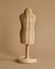 In a cozy dressing room, the Minikane Mannequin For Doll Clothing stands against a beige background. With its woven texture and circular wooden base, this wicker dress form resembles a mannequin and adds to the minimalist and rustic ambiance. The neutral tones of both the dress form and the background create a serene scene.