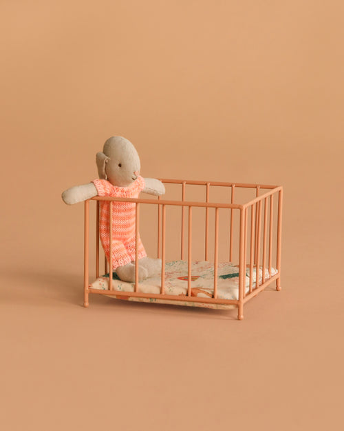 A small stuffed doll in a pink outfit stands inside the Maileg Playpen, Micro - Dark powder, featuring a patterned mattress and set against a plain peach background.